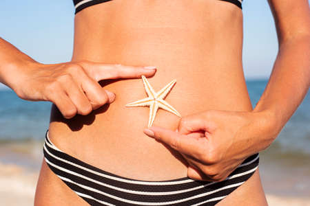 Woman on the beach in a swimsuit holding a starfish on the stomach.の写真素材