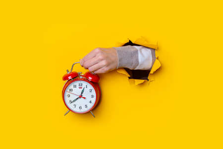 Female hand holding a red watch in a hole on a yellow background.