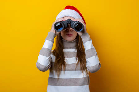 Young woman wearing a Santa Claus hat looks through binoculars on a yellow background.の写真素材