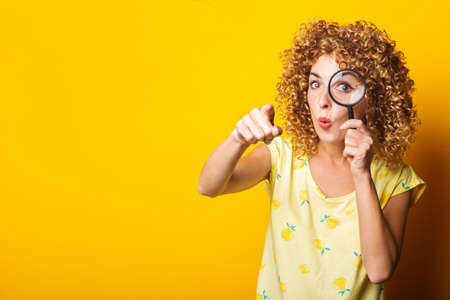 curly girl pointing the finger in front of a looking through a magnifying glass on a yellow...