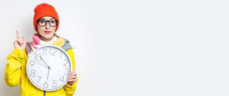 Young woman pointing her finger up while holding a clock on a light background. Banner