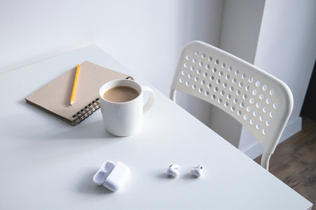 White cup with coffee, notepad, pencil and wireless headphones on a white table