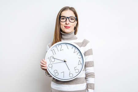 Smiling young woman in glasses holding white wall clock on light background