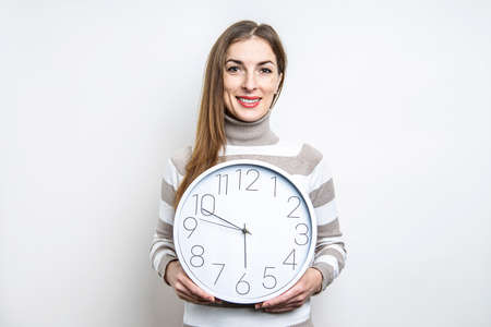 Smiling young woman holding white wall clock on light background