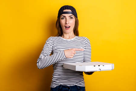surprised young woman pizza delivery man, points finger to the side while holding package with hot pizza on yellow background.の写真素材