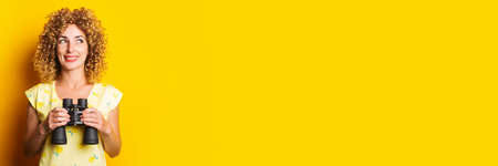 smiling beautiful curly young woman with binoculars on yellow background. Bannerの写真素材