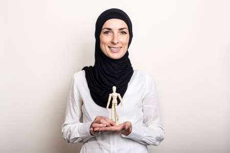 A smiling young woman in a hijab holds a puppet of a wooden man on a light background.