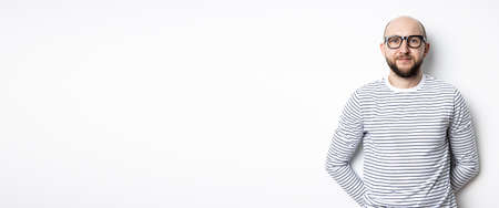 Bearded man with glasses leaned against a white wall.