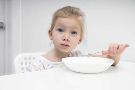Sad child girl blonde eats porridge at a white tableの写真素材