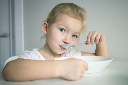 Child girl blonde eats with a spoon from a white plate at a white tableの写真素材