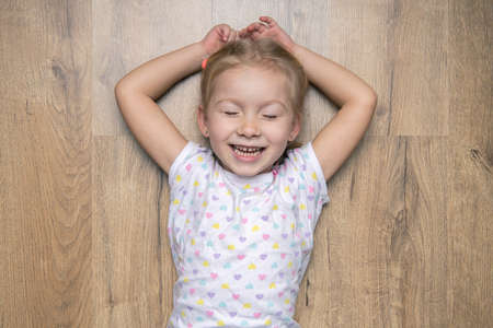 Smiling child girl with closed eyes lying on a wooden floor. Top view, flat lay