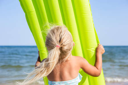 Blond child girl in a swimsuit holding an inflatable mattress on the beach