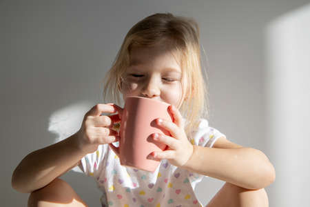 Child girl blonde with closed eyes holds a cup on the background of a white wallの写真素材