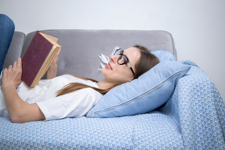 A young woman in glasses lies on a sofa reading a book.の写真素材