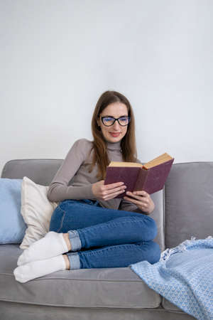 Smiling young woman in glasses sitting on sofa reading a book.