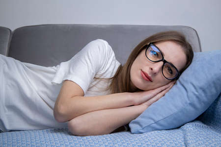 Serious young woman in white t-shirt with glasses lying on sofa.