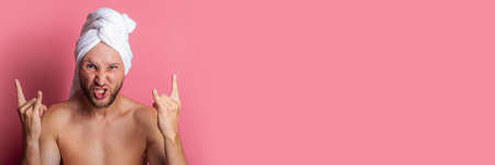 bearded young man with a towel on his head shows the gesture of a goat on a pink background. Banner