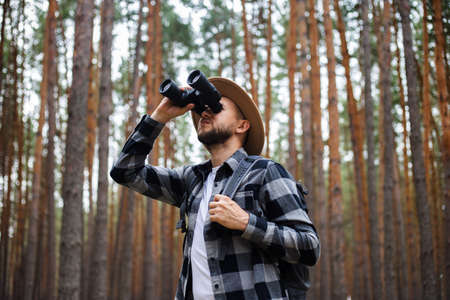 Young Man in a hat with a backpack and binoculars in a pine forest. Hike in the mountains or forest.の写真素材