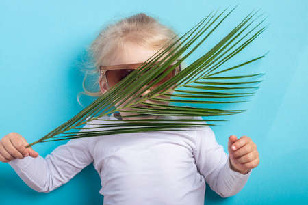Beautiful blonde girl with a palm leaf in sunglasses lying on a blue background. Top view, flat lay.