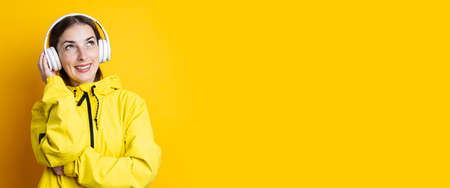 Smiling young woman in headphones in a yellow jacket looks up on a yellow background. Banner
