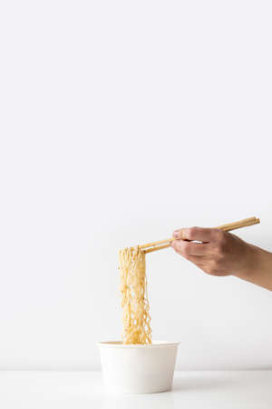 Female hand takes Chinese noodles with Chinese chopsticks from a paper bowl on a white background.