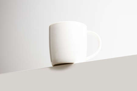 White cup stands on the edge of the table on a white background.