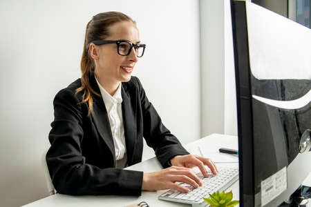 Smiling young woman in glasses working while sitting at a computer in the workplace.の写真素材