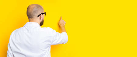 Young man standing with his back pointing his finger at an invisible screen on a yellow background. Banner.の写真素材