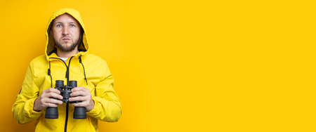 Young man in a yellow jacket holding binoculars on a yellow background. Banner.の写真素材