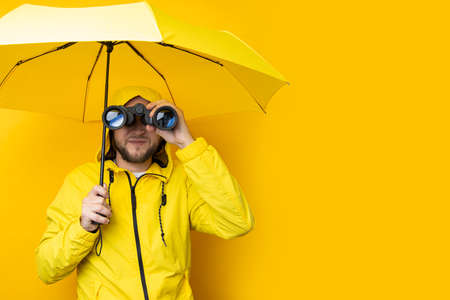 Young man in a raincoat with an umbrella looks through binoculars on a yellow background.の写真素材