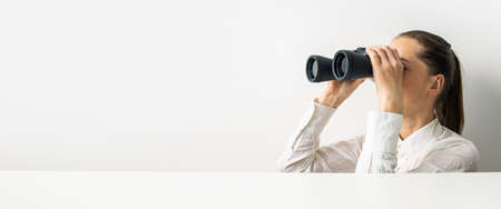 Young woman in shirt looking through binoculars looking out from behind cardboard. Banner.