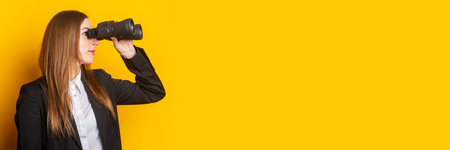 young woman in a jacket looks through binoculars on a yellow background. Banner.
