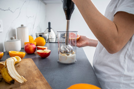 Woman using hand blender to mix fresh fruit smoothie in kitchen.の写真素材
