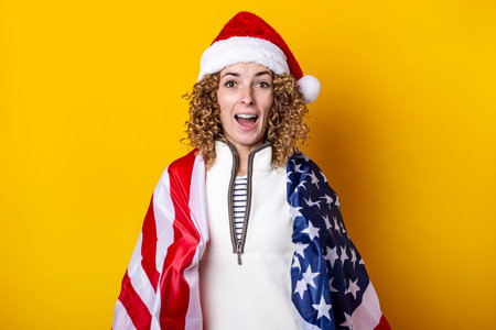 Young woman in santa claus hat shouting joyfully with the usa flag on a yellow backgroundの写真素材