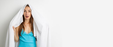 Surprised young woman covering her head with white sheet on white background. Banner.