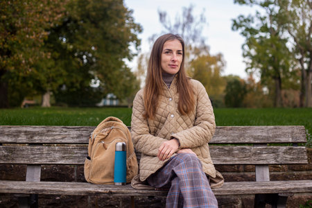 Smiling young woman with thermos sitting on park bench.の写真素材
