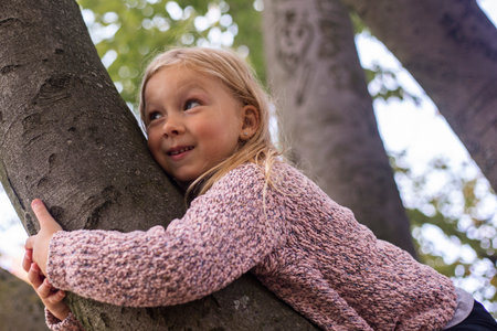 Smiling blond girl child lies hugging a tree with two hands.の写真素材