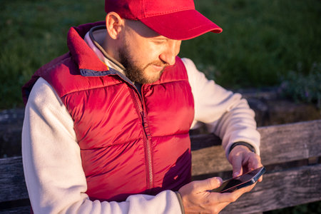 Young man with a beard with a phone sitting on a park bench.の写真素材
