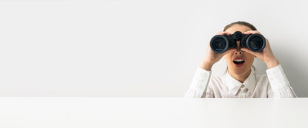 Surprised woman looking through binoculars peeking out from behind white cardboard. Banner.の写真素材