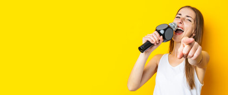 Singing young woman holding microphone pointing finger forward on yellow background. Banner.の写真素材