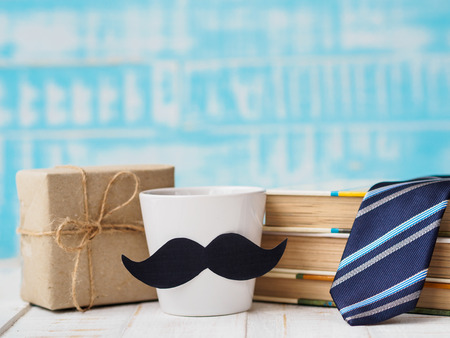 Happy father's day concept. Gift box, a cup of coffee with mustache, stack of books and necktie on blue wooden texture background.の写真素材