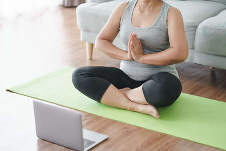 Mature Asian chubby fat woman sitting on the floor in living room practice online yoga lesson with the computer. female having meditate training class on the laptop.の写真素材