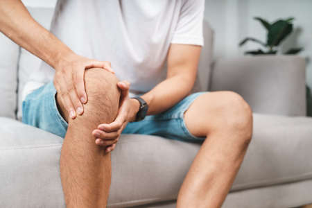 The man suffering from knee pain sitting on the couch hold and massaging his painful knee.の写真素材