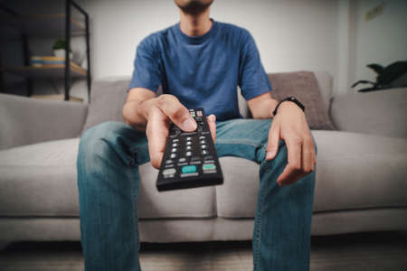 Man hold and press the TV remote controller button sitting on the couch.