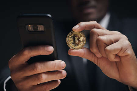 Businessman in black suit holding a golden bitcoin using Smartphone with Bitcoin trading chart on...