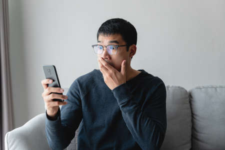 Portrait of excited asian man looking at mobile phone, man receiving good news using smartphone at home.の写真素材