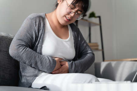 Woman suffering from stomachache sitting on the sofa at home. abdominal pain, Gastritis, Period,...