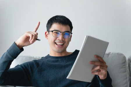 Young thoughtful Asian man in glasses thinking and looking at notebook.