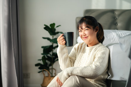Young woman drinking coffee in bedroom, Holding coffee cup with two handsの写真素材