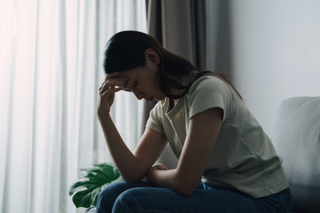 Unhappy lonely depressed woman is sitting on the couch and looking down. She is wearing a white shirt and blue jeansの写真素材